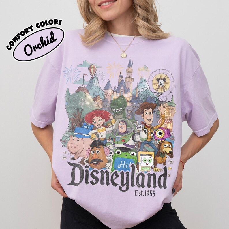 May include: A light purple t-shirt featuring a graphic print of various Toy Story characters in front of a Disneyland castle. The text reads Disneyland Est. 1955. A small oval label in the top left corner displays the text Comfort Colors Orchid.