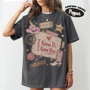 Puede incluir: Una camiseta gris carb&oacute;n de manga corta con un estampado gr&aacute;fico de una nota de papel rayado con el texto I Knew It, I Knew You, rodeado de ilustraciones de un sombrero de vaquero rosa, una estrella de sheriff, una rueda y una bota.
