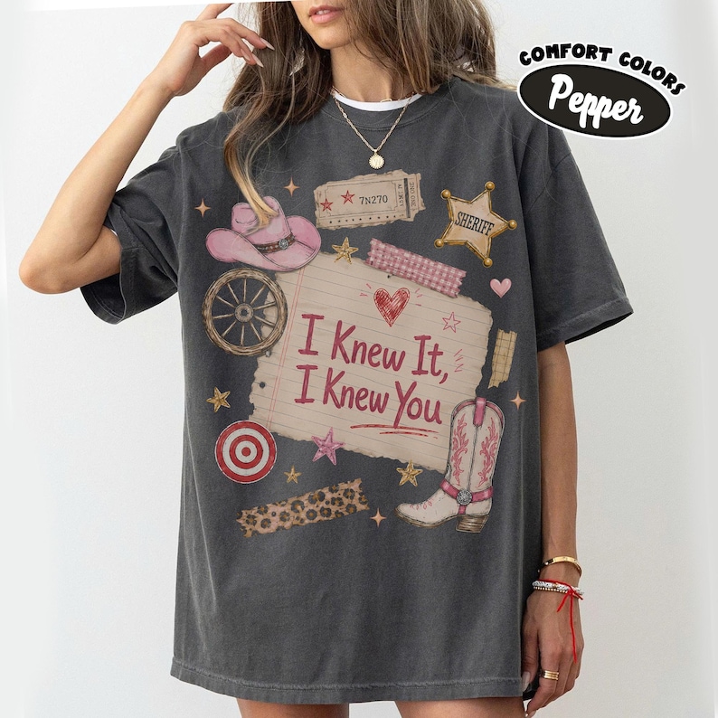 Puede incluir: Una camiseta gris carb&oacute;n de manga corta con un estampado gr&aacute;fico de una nota de papel rayado con el texto I Knew It, I Knew You, rodeado de ilustraciones de un sombrero de vaquero rosa, una estrella de sheriff, una rueda y una bota.