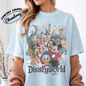 May include: Light blue t-shirt featuring a colorful graphic of Disney characters in front of a castle and amusement park. The word "Disneyworld" is printed in a vintage font, with "Est.1971" below. The shirt has the words "Comfort Colors" and "Chambray" printed on the top left.