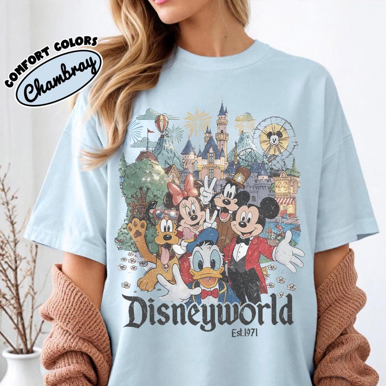 May include: Light blue t-shirt featuring a colorful graphic of Disney characters in front of a castle and amusement park. The word "Disneyworld" is printed in a vintage font, with "Est.1971" below. The shirt has the words "Comfort Colors" and "Chambray" printed on the top left.