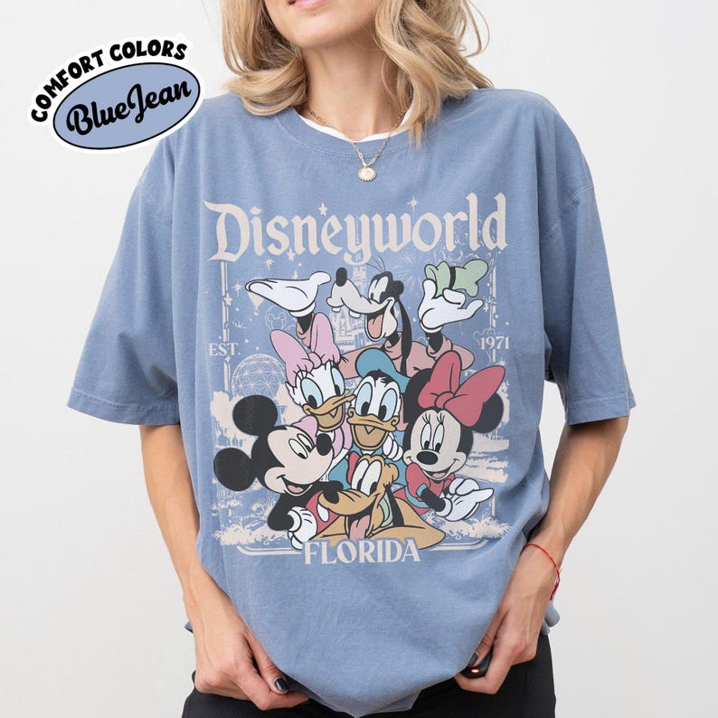 May include: Blue t-shirt with a Disney World graphic featuring Mickey Mouse, Minnie Mouse, Donald Duck, Daisy Duck, Goofy, and Pluto. The shirt has the text "Disneyworld Florida" and "EST. 1971". The shirt is a Comfort Colors Blue Jean.