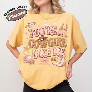 Puede incluir: Una camiseta de color amarillo mostaza con un estampado gr&aacute;fico que incluye el texto "YOU'RE A COWGIRL LIKE ME" en rosa y marr&oacute;n. El dise&ntilde;o presenta un cohete, un sombrero de vaquero, una herradura, una estrella de sheriff, una bola de discoteca, un lazo y botas.