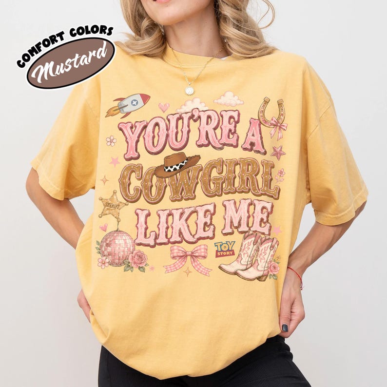 Puede incluir: Una camiseta de color amarillo mostaza con un estampado gr&aacute;fico que incluye el texto "YOU'RE A COWGIRL LIKE ME" en rosa y marr&oacute;n. El dise&ntilde;o presenta un cohete, un sombrero de vaquero, una herradura, una estrella de sheriff, una bola de discoteca, un lazo y botas.