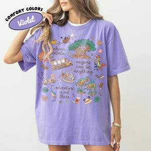 Puede incluir: Una camiseta violeta Comfort Colors con un dise&ntilde;o gr&aacute;fico colorido. El dise&ntilde;o incluye los textos "Hakuna Matata", "Anyone can be anything" y "Adventure is out there", junto con ilustraciones de dibujos animados.