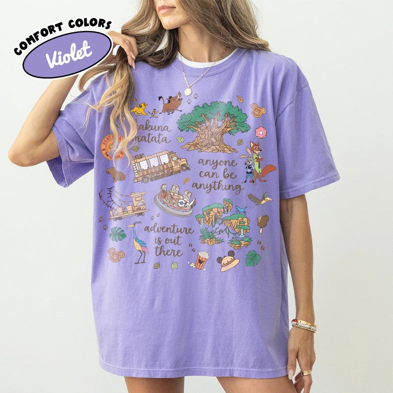 Puede incluir: Una camiseta violeta Comfort Colors con un dise&ntilde;o gr&aacute;fico colorido. El dise&ntilde;o incluye los textos "Hakuna Matata", "Anyone can be anything" y "Adventure is out there", junto con ilustraciones de dibujos animados.
