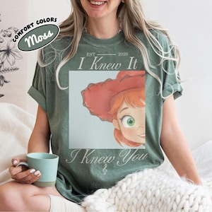 Op de afbeelding: Een mosgroen T-shirt met korte mouwen en een grafische print van een stripfiguur met rood haar en een rode hoed. De tekst luidt EST 2026 I Knew It I Knew You. Een klein label in de hoek toont de tekst COMFORT COLORS MOSS.
