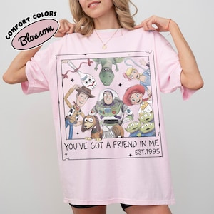 May include: A light pink t-shirt featuring a framed graphic of Toy Story characters, including Woody, Buzz Lightyear, and others. The text "YOU'VE GOT A FRIEND IN ME" and "EST.1995" are printed below the graphic. The shirt has the words "COMFORT COLORS" and "Blossom" printed on the top left.