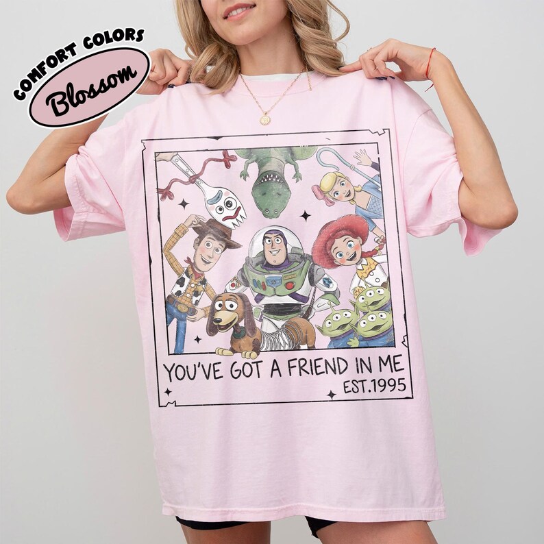 May include: A light pink t-shirt featuring a framed graphic of Toy Story characters, including Woody, Buzz Lightyear, and others. The text "YOU'VE GOT A FRIEND IN ME" and "EST.1995" are printed below the graphic. The shirt has the words "COMFORT COLORS" and "Blossom" printed on the top left.