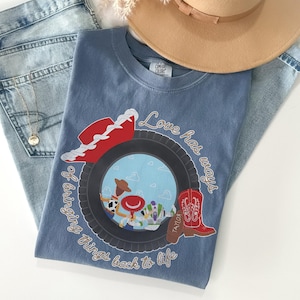 May include: A blue t-shirt featuring a graphic of a black tire framing a scene with three cartoon characters, a red cowboy hat, and a red cowboy boot. The text "Love has ways of bringing things back to life" is printed in a cursive font around the tire.