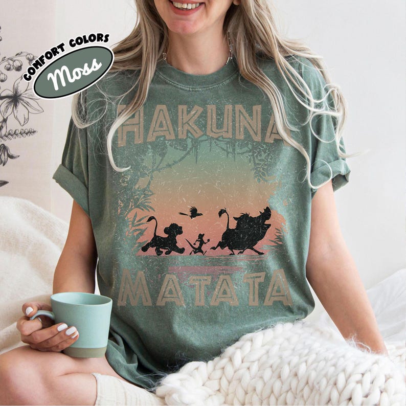 May include: A moss green t-shirt with a graphic featuring the words "HAKUNA MATATA" and silhouettes of characters from a popular animated film. The shirt has rolled-up sleeves and a relaxed fit. The words "COMFORT COLORS" and "Moss" are in an oval.