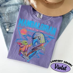 Może przedstawiać: Fioletowa koszulka Comfort Colors z grafiką The Mandalorian i Grogu. Projekt zawiera tekst "MANDALORIAN" i "THIS IS THE WAY" w kolorze niebieskim. Beżowy kapelusz i dżinsy są w tle.