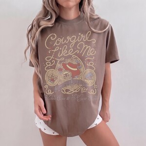 May include: A brown short-sleeve t-shirt featuring a graphic print with the text "Cowgirl Like Me" and "Takes One to Know One". The design includes a red cowboy hat, a horseshoe, a bull skull, and rope motifs in a light yellow and red color palette.