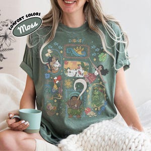May include: A moss green t-shirt with Disney characters and the word "Disney" at the top. The shirt has a crew neck and short sleeves. The words "Comfort Colors" and "Moss" are printed on a label in the upper left corner.
