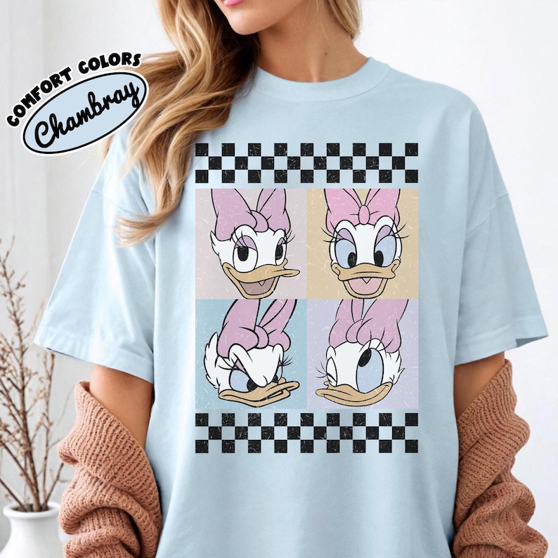 May include: Light blue t-shirt featuring a four-panel Daisy Duck graphic with pink bows and a checkered border. The shirt includes the text "COMFORT COLORS Chambray" in a circular design.