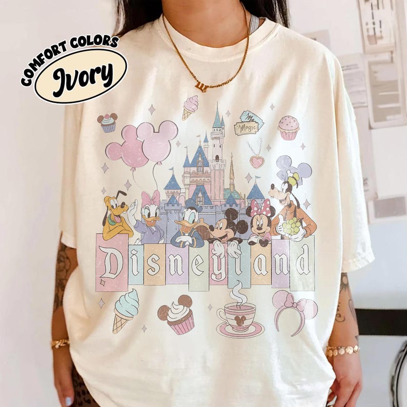 May include: An ivory t-shirt featuring a graphic print of a castle, Mickey Mouse, Minnie Mouse, Donald Duck, Daisy Duck, Goofy, and Pluto. The design includes pastel-colored balloons, cupcakes, a coffee cup, and the text Disneyland in a serif font.