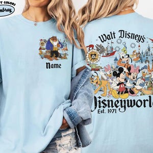 May include: Light blue t-shirt featuring a colorful graphic of Disney characters and the text "Walt Disney's Disneyworld Est. 1971" on the back. The front of the shirt has a graphic of the Beast and the word "Name".