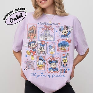 May include: A light purple t-shirt with a Walt Disney World design. The shirt features various Disney characters and landmarks in a postage stamp style, with the text "250 years of freedom" and "2026". The shirt is labeled "Comfort Colors Orchid".