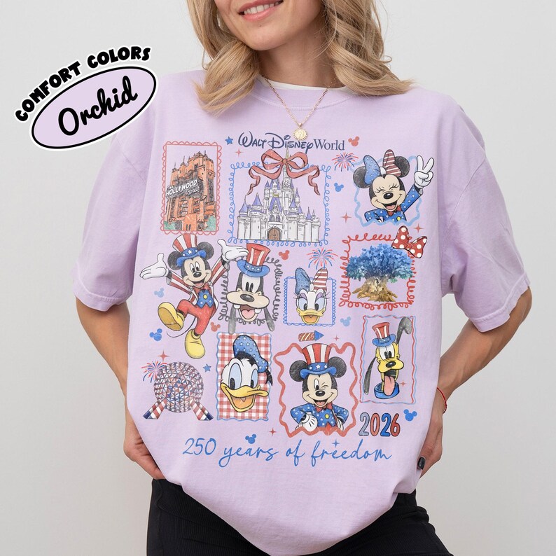 May include: A light purple t-shirt with a Walt Disney World design. The shirt features various Disney characters and landmarks in a postage stamp style, with the text "250 years of freedom" and "2026". The shirt is labeled "Comfort Colors Orchid".