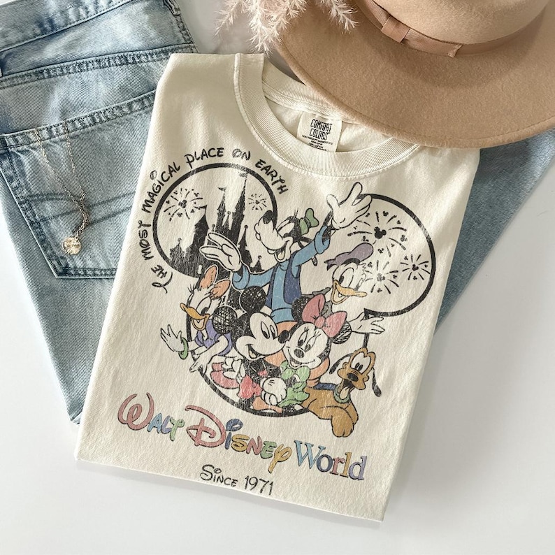 May include: Cream-colored Walt Disney World t-shirt featuring Mickey Mouse, Minnie Mouse, Donald Duck, Daisy Duck, Goofy, and Pluto. The shirt has the text "The Most Magical Place on Earth" and "Since 1971". The shirt is folded on top of blue jeans.
