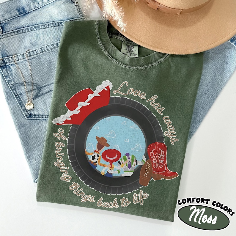 May include: An olive green crew neck t-shirt featuring a graphic of a black tire framing a blue sky with cartoon characters. The design includes a red cowboy hat, a red cowboy boot labeled Taylor, and the text Love has ways of bringing things back to life.