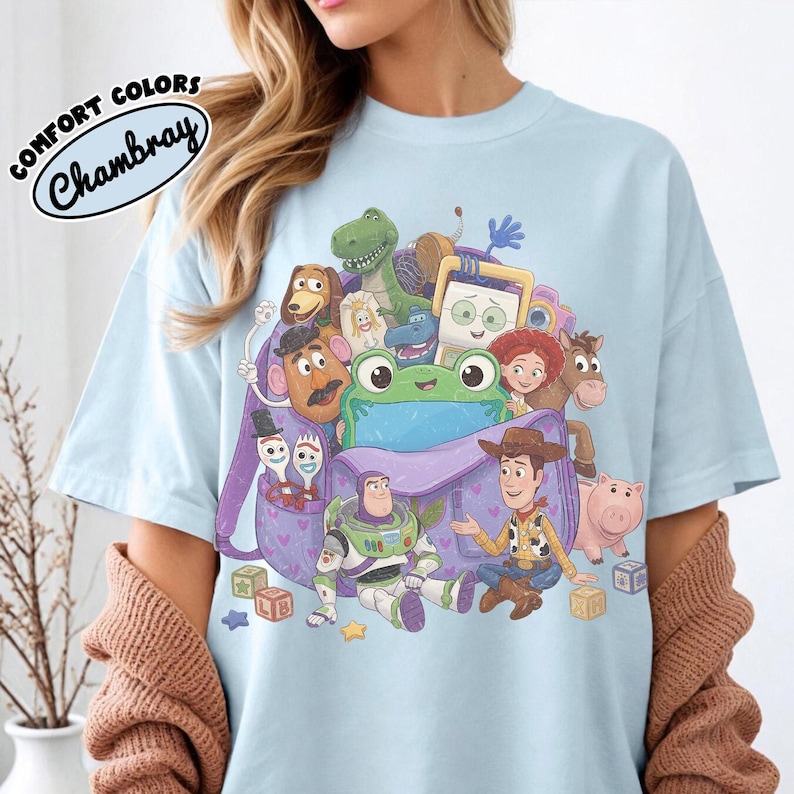 Pode incluir: Uma t-shirt azul claro com uma estampa gr&aacute;fica de v&aacute;rios personagens animados de Toy Story, incluindo Woody, Buzz Lightyear e um sapo verde, reunidos em torno de uma mochila roxa. O texto Comfort Colors Chambray &eacute; vis&iacute;vel.