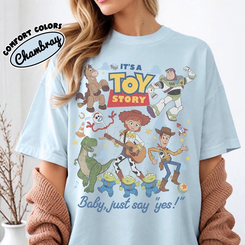 May include: A light blue t-shirt featuring a graphic print of Toy Story characters including Woody, Jessie, Buzz Lightyear, Rex, Bullseye, Forky, and three aliens. The text reads IT'S A TOY STORY and Baby, just say yes! with a Comfort Colors logo.