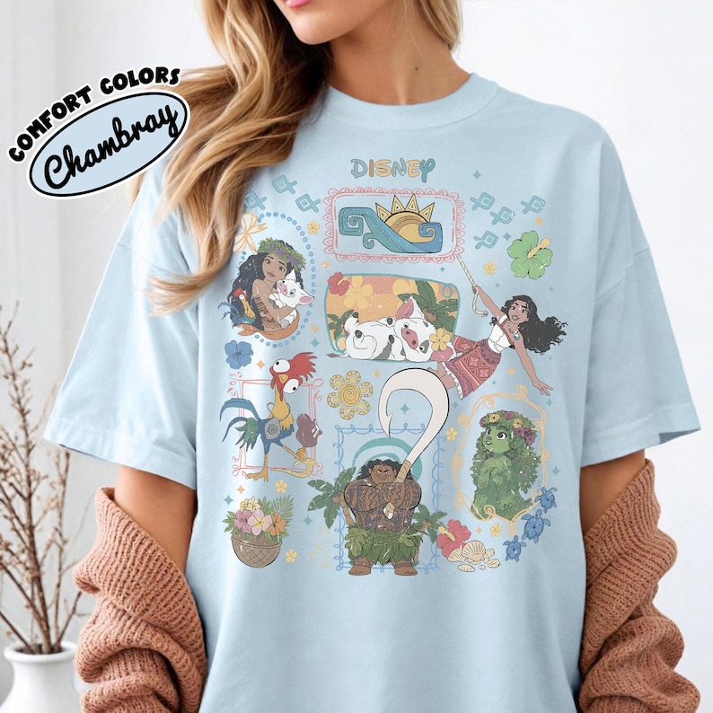 May include: Pale blue t-shirt with a Disney design featuring characters from the film Moana. The shirt has a crew neck and short sleeves. The design includes illustrations of Moana, Maui, Pua, Hei Hei, and other characters, with the word "Disney" printed at the top.