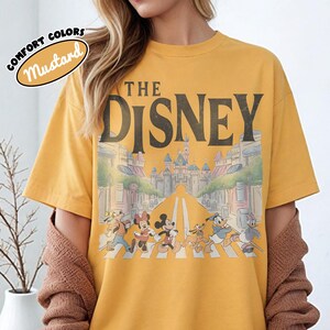 Może przedstawiać: Musztardowa koszulka z napisem "THE DISNEY" nad grafiką postaci Disneya przechodzących przez ulicę. Grafika zawiera Myszkę Miki, Minnie, Kaczora Donalda i Goofy'ego. Koszulka ma okrągły dekolt i kr&oacute;tkie rękawy.