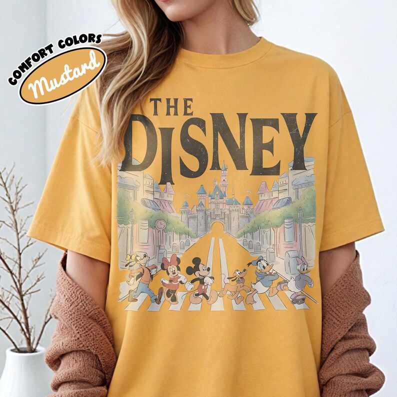 Może przedstawiać: Musztardowa koszulka z napisem "THE DISNEY" nad grafiką postaci Disneya przechodzących przez ulicę. Grafika zawiera Myszkę Miki, Minnie, Kaczora Donalda i Goofy'ego. Koszulka ma okrągły dekolt i kr&oacute;tkie rękawy.