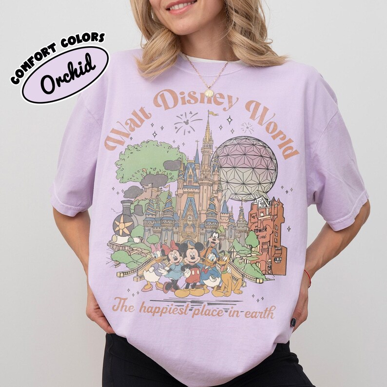May include: A light purple t-shirt with a graphic of the Walt Disney World logo and characters. The shirt has the words "Walt Disney World" and "The happiest place on earth" printed on it. The shirt also has the words "Comfort Colors" and "Orchid" printed on it.