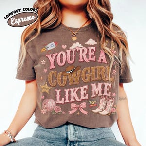 Puede incluir: Una camiseta de manga corta de color marr&oacute;n oscuro con el texto "YOU'RE A COWGIRL LIKE ME" en rosa y marr&oacute;n. El gr&aacute;fico incluye un cohete, un sombrero de vaquero, una estrella de sheriff, una bola de discoteca, un lazo rosa y botas con el logo "TOY STORY".