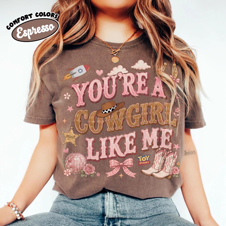 Puede incluir: Una camiseta de manga corta de color marr&oacute;n oscuro con el texto "YOU'RE A COWGIRL LIKE ME" en rosa y marr&oacute;n. El gr&aacute;fico incluye un cohete, un sombrero de vaquero, una estrella de sheriff, una bola de discoteca, un lazo rosa y botas con el logo "TOY STORY".