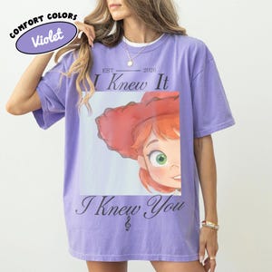 Op de afbeelding: Een violetkleurig T-shirt met korte mouwen en een grafische print van een stripfiguur met rood haar en een rode hoed. De tekst luidt "EST 2026", "I Knew It" en "I Knew You" met een klein muzieknootsymbool. Het shirt heeft een ronde hals.