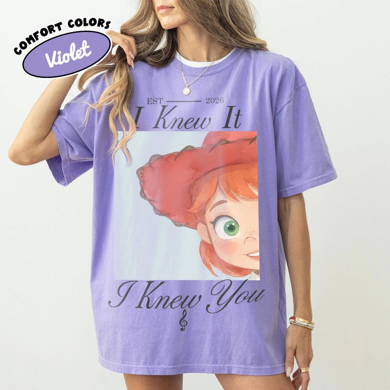 Op de afbeelding: Een violetkleurig T-shirt met korte mouwen en een grafische print van een stripfiguur met rood haar en een rode hoed. De tekst luidt "EST 2026", "I Knew It" en "I Knew You" met een klein muzieknootsymbool. Het shirt heeft een ronde hals.