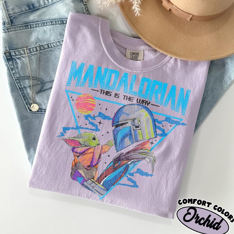 Może przedstawiać: Lawendowa koszulka Comfort Colors z grafiką Mandalorianina i Grogu oraz napisem "MANDALORIAN - THIS IS THE WAY." Projekt zawiera r&oacute;żowo-ż&oacute;łtą planetę i niebieski tr&oacute;jkąt. Koszulka jest złożona na niebieskich dżinsach.