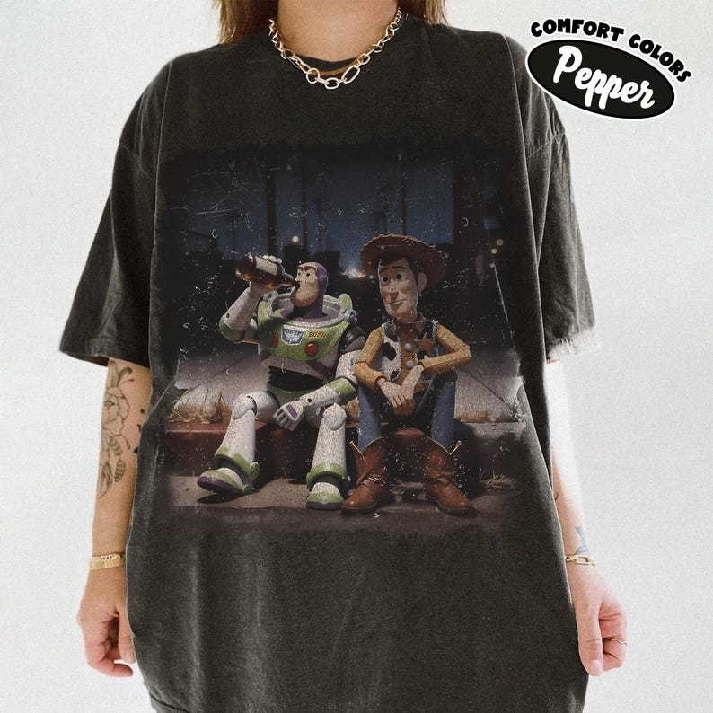 May include: A charcoal gray short-sleeve t-shirt featuring a graphic print of Buzz Lightyear and Woody sitting together. The graphic shows Buzz holding a bottle and Woody sitting beside him. A Comfort Colors Pepper label is visible in the top right corner.