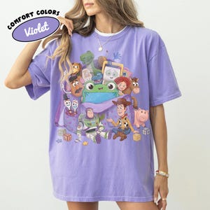 Pode incluir: Uma t-shirt violeta de manga curta com uma impress&atilde;o gr&aacute;fica de v&aacute;rias personagens animadas, incluindo um cowboy, um ranger espacial, um sapo verde e um porco. O texto COMFORT COLORS Violet est&aacute; impresso no canto superior esquerdo.