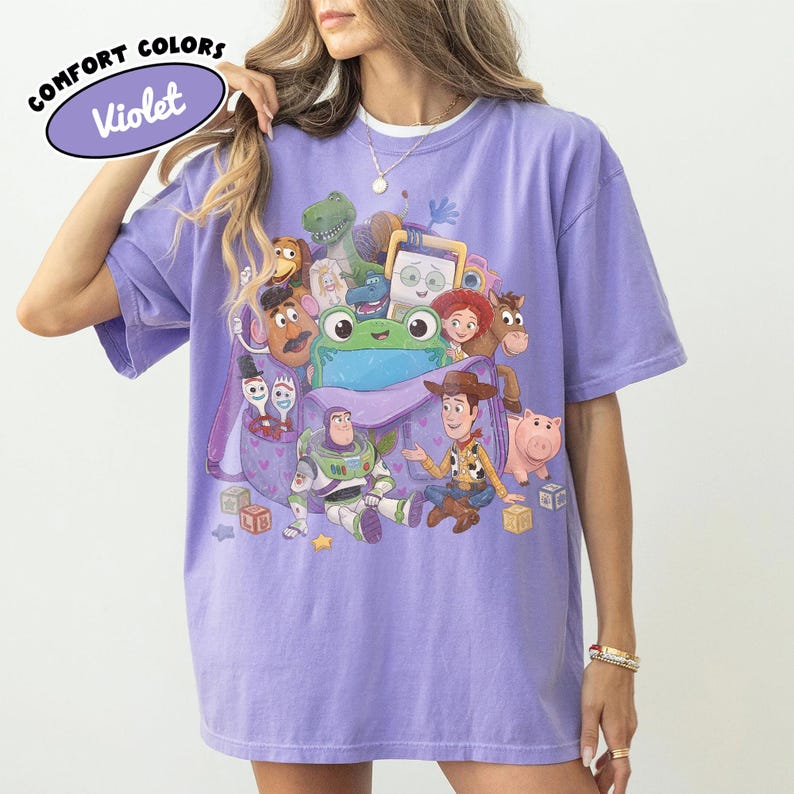 Pode incluir: Uma t-shirt violeta de manga curta com uma impress&atilde;o gr&aacute;fica de v&aacute;rias personagens animadas, incluindo um cowboy, um ranger espacial, um sapo verde e um porco. O texto COMFORT COLORS Violet est&aacute; impresso no canto superior esquerdo.