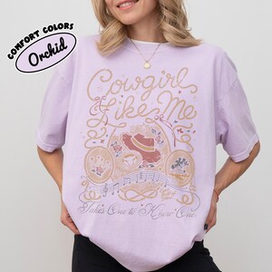 May include: A light purple t-shirt featuring a graphic design with the text "Cowgirl Like Me" and "Takes One to Know One". The illustration includes a cowboy hat, a horseshoe, musical notes, and rope motifs in shades of brown and cream.