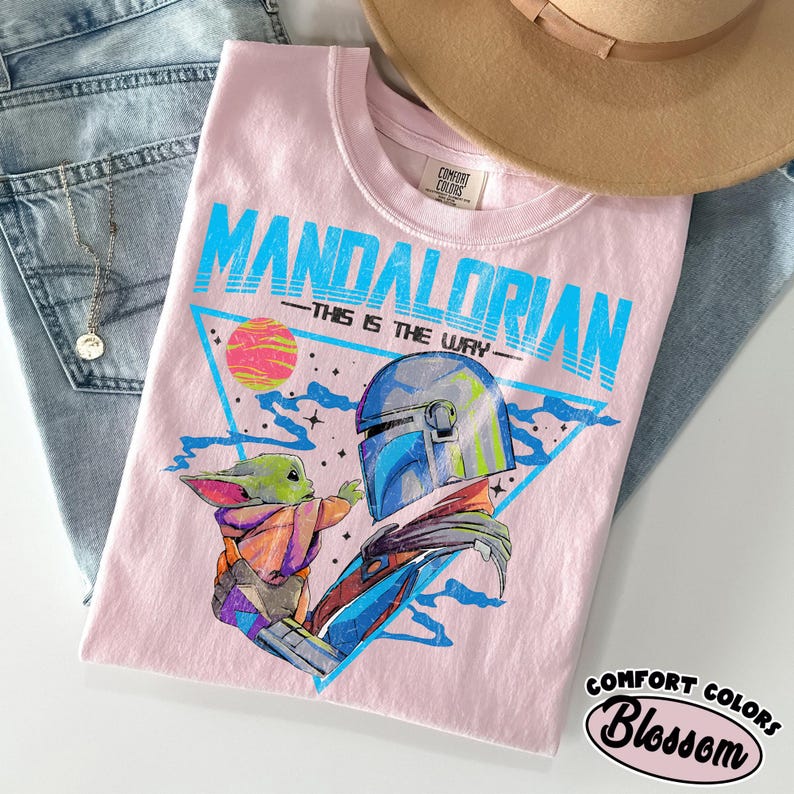 Może przedstawiać: R&oacute;żowa koszulka z grafiką Mandalorianina i Grogu, z niebieskim napisem "MANDALORIAN - THIS IS THE WAY". Koszulka to Comfort Colors Blossom, z okrągłym dekoltem.
