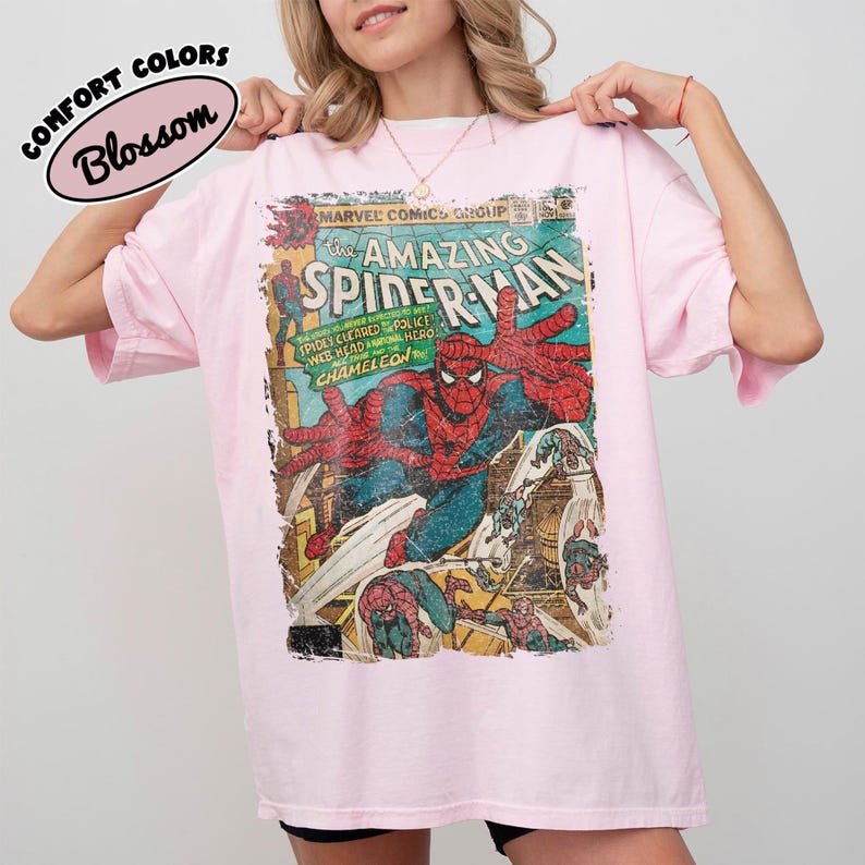 May include: A light pink short-sleeve t-shirt featuring a vintage-style graphic of The Amazing Spider-Man comic book cover. The print displays Spider-Man in a red and blue suit with text including Marvel Comics Group and Chameleon.