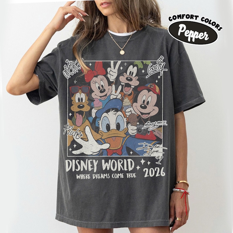 May include: Dark gray t-shirt featuring a graphic of Disney characters including Mickey Mouse, Donald Duck, and Goofy. The graphic is framed with the words "DISNEY WORLD 2026" and "WHERE DREAMS COME TRUE". The shirt also has the words "COMFORT COLORS Pepper".
