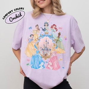 May include: Orchid Comfort Colors t-shirt featuring a colorful illustration of several princesses around a castle. The princesses are wearing gowns in shades of blue, yellow, pink, and green. The shirt has the words "COMFORT COLORS" and "Orchid" printed on it.