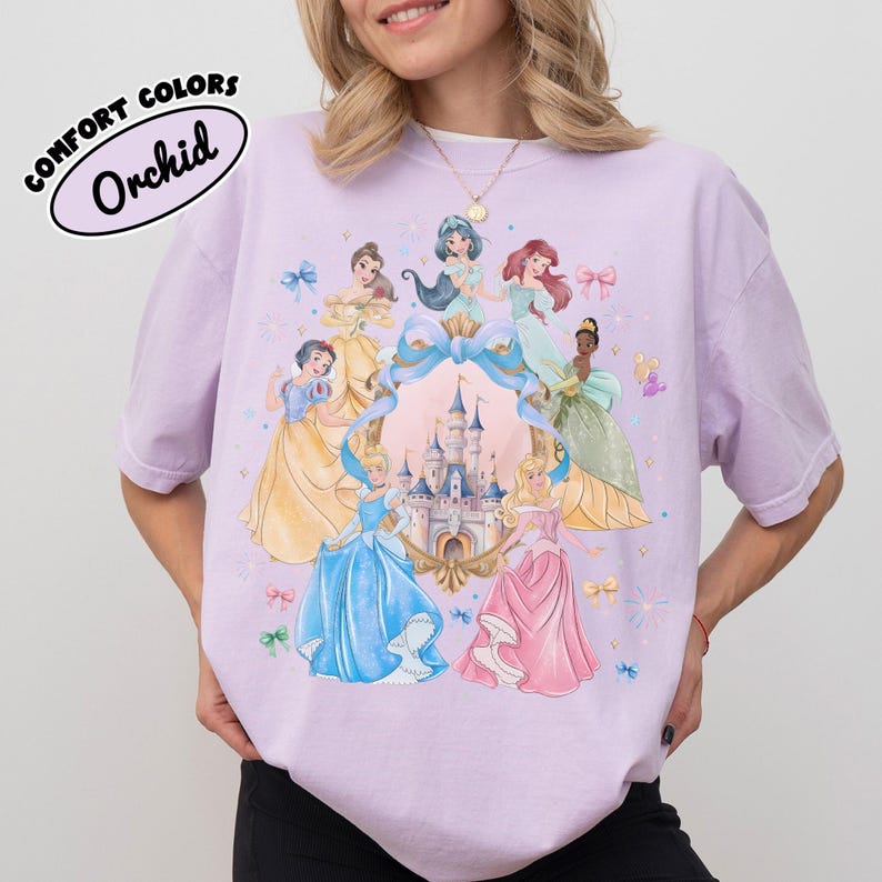 May include: Orchid Comfort Colors t-shirt featuring a colorful illustration of several princesses around a castle. The princesses are wearing gowns in shades of blue, yellow, pink, and green. The shirt has the words "COMFORT COLORS" and "Orchid" printed on it.