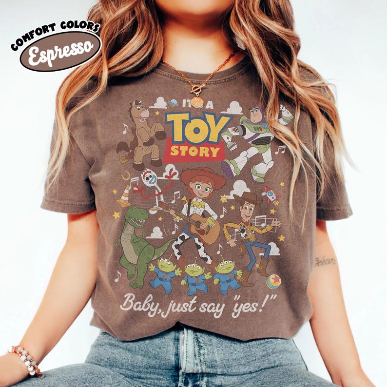 May include: A dark brown short-sleeve t-shirt featuring a graphic print of Toy Story characters including Woody, Jessie, Buzz Lightyear, Rex, Forky, Bullseye, and three aliens. The text reads IT'S A TOY STORY and Baby, just say yes!