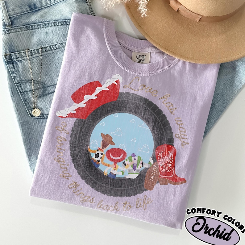 May include: A light purple t-shirt featuring a graphic of a tire framing a blue sky scene with cartoon characters. A red cowboy hat and a red cowboy boot are positioned near the tire. The text reads Love has ways of bringing things back to life.