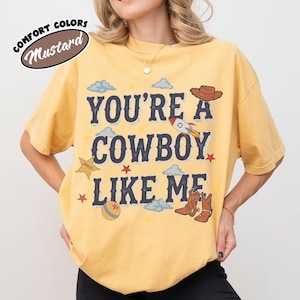 May include: A mustard yellow short-sleeve t-shirt featuring the dark blue text "YOU'RE A COWBOY LIKE ME" surrounded by graphic illustrations of a brown cowboy hat, a rocket ship, a sheriff star, a ball, and a pair of brown cowboy boots.