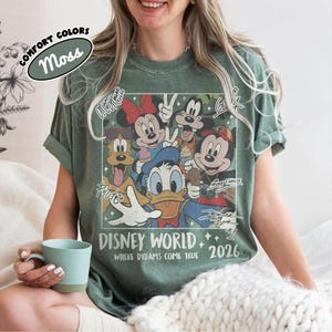 May include: Moss green t-shirt featuring a Disney World graphic with Mickey Mouse, Minnie Mouse, Donald Duck, and other characters. The shirt includes the text "Disney World Where Dreams Come True 2026".