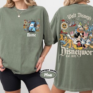 May include: Olive green t-shirt featuring a cartoon Mater design with the text "Name" on the front. The back of the shirt displays a colorful Disney World graphic with various characters and the text "Walt Disney's Disneyworld Est. 1971".